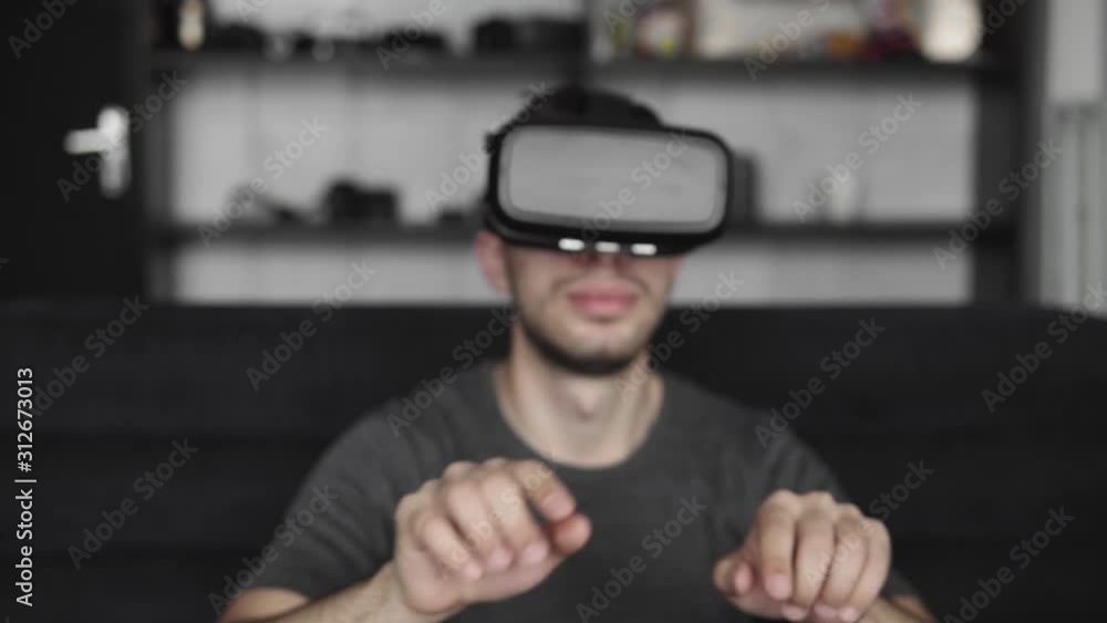 Young bearded man wearing headset of virtual reality in office sitting on a sofa and trying to touch something that he see in virtual world. Using VR headset for work. Young bearded man wearing headset of virtual reality in office sitting on a sofa and trying to touch something that he see in virtual world. Using VR headset for work.