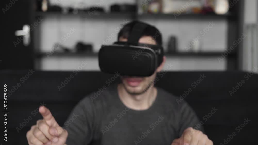 Young bearded man wearing headset of virtual reality in office sitting on a sofa and trying to touch something that he see in virtual world. Using VR headset for work. Young bearded man wearing headset of virtual reality in office sitting on a sofa and trying to touch something that he see in virtual world. Using VR headset for work.