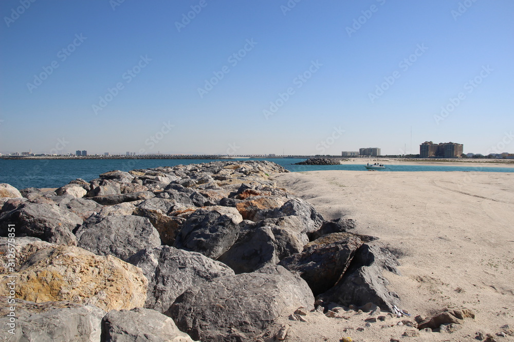 Big stones on the sea coast with white sand beach, bay, sea, buildings on horizon and blue sky