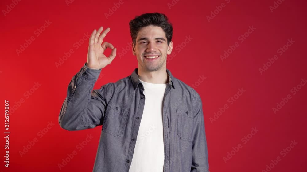 Handsome young man in denim shirt smiles to camera. Hipster guy showing OK sign over red background. Winner. Success. Body language. Handsome young man in denim shirt smiles to camera. Hipster guy showing OK sign over red background. Winner. Success. Body language.