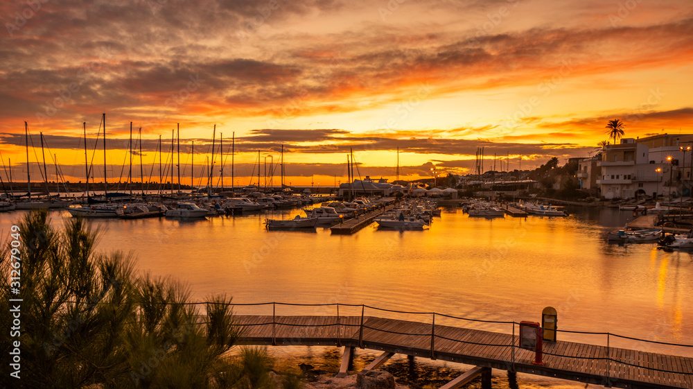 A view of the marina of Santa Maria di Leuca at red and orange sunset. Sailboats and yachts moored on the docks of the port. Wooden walkways lead to harbor quay. Puglia, Italy, Lecce, Salento.