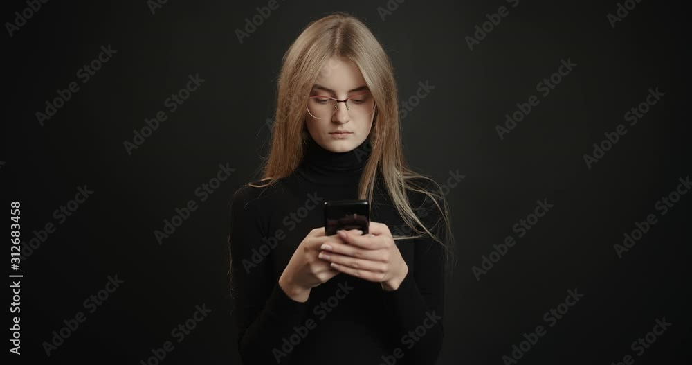 Focused blonde woman in black outfit and eyeglasses using smartphone, isolated on dark background, technologies Focused blonde woman in black outfit and eyeglasses using smartphone, isolated on dark background, technologies