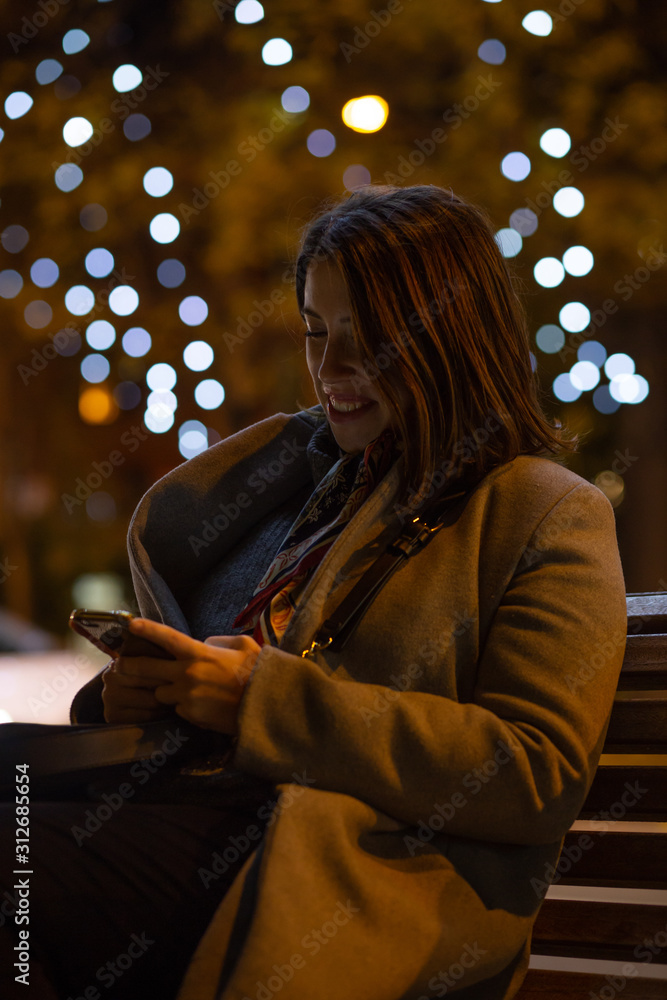 young woman sitting on a bench while looking at her phone at night