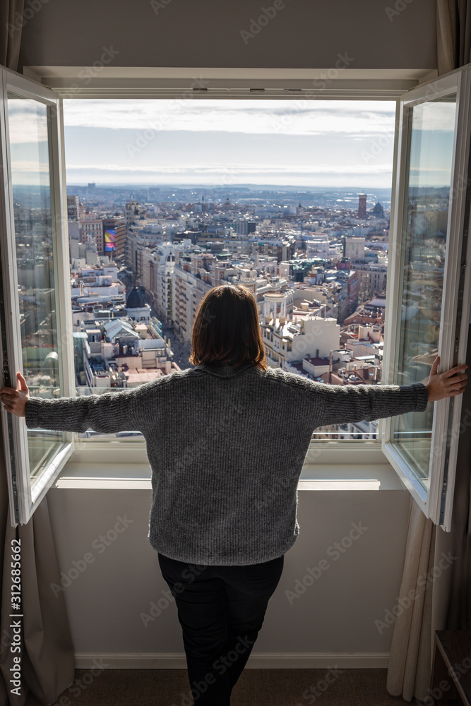 Obraz premium rear view of young woman opening a window from her room and looking outside on a sunny day with Madrid skyline views
