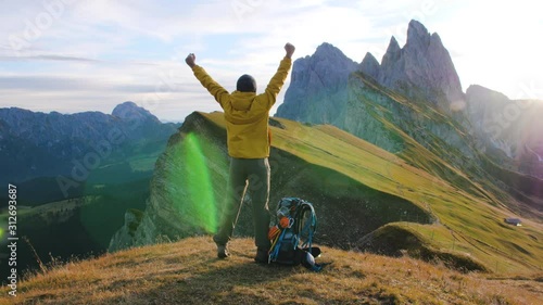 Young man hiking at Seceda mountain raising arms to the sky on the peak at sunrise. Backpack, yellow jacket, boots, beanie. Traveling to puez Odle, Dolomites, Trentino, Italy.
