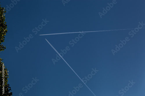 Two airplanes with jet stream in a clear blue sky not about to collide but pass without danger above the French town of Giverny framed by a green shrub on the left.