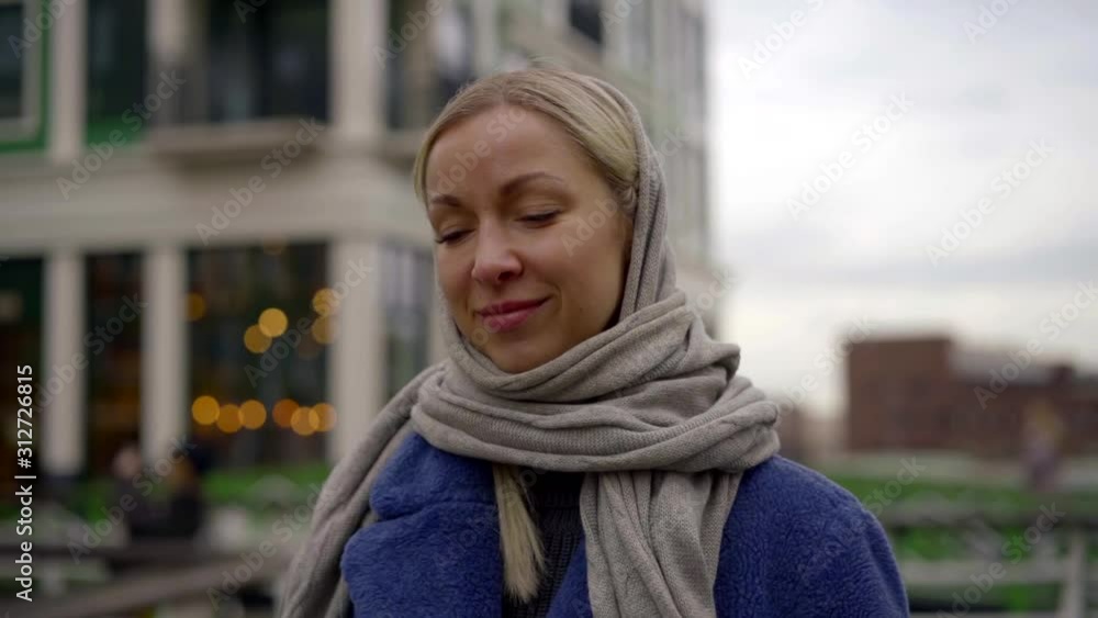A close-up portrait of a fair-haired girl with a headscarf on her head, wearing a blue coat, she is against the background of European houses and the river. A close-up portrait of a fair-haired girl with a headscarf on her head, wearing a blue coat, she is against the background of European houses and the river.