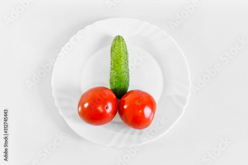 Red ripe juicy tomatoes and green cucumber on a white plate. The phallic image of natural components. Light gray background photo