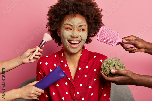 Sincere glad woman applies clay facial mask, turns gaze aside, wears red pajama, gets various beauty treatments at same time, surrounded with hair comb, cream and makeup brush, isolated over rosy wall