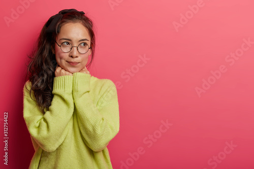 Brunette millennial young Asian woman keeps hands under chin, wears transparent glasses and green jumper, thinks deeply about something, isolated on pink wall with copy space area. People and thoughts
