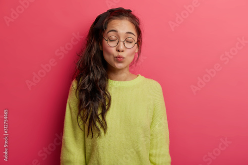 Horizontal shot of lovely brunette Asian woman with lips rounded, keeps eyes shut, has pony tail, wears optical round glasses and green jumper, isolated over rosy wall, gives mwah and sends kiss