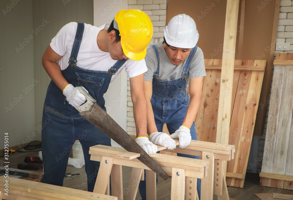 Two Asian repairmen wearing safety helmet , working in wood workshop , cutting wooden plank with hand saw. Two Asian repairmen wearing safety helmet , working in wood workshop , cutting wooden plank with hand saw.