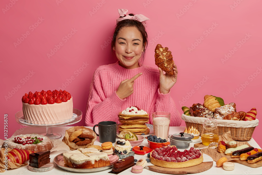 Beautiful smiling Asian woman points at delicious croissant, wears knitted sweater, sits at festive table with tasty sweet desserts, drinks fresh milk, cannot imagine her life without sugar. Beautiful smiling Asian woman points at delicious croissant, wears knitted sweater, sits at festive table with tasty sweet desserts, drinks fresh milk, cannot imagine her life without sugar.