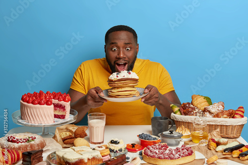 Dark skinned man tempted by delicious creamy pancakes, opens mouth, wears yellow t shirt, has sweet tooth, sits at table with various desserts, isolated over blue background. Pastry, bakery.