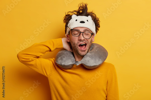 Sleepy male traveler yawns as wants to sleep, travels in transport with neck pillow, wears transparent glasses, headband, yellow jumper, poses indoor. People, tiredness and travelling concept photo