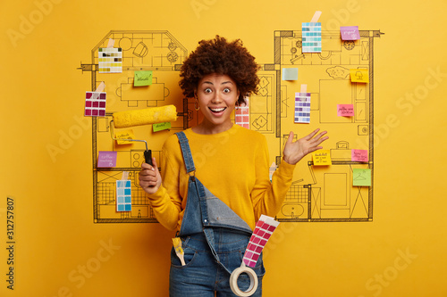 Horizontal shot of happy Afro woman paints walls at home, holds yellow paint roller, has positive look, busy with refurbishing house, wears denim dungarees, stands over architectural project photo