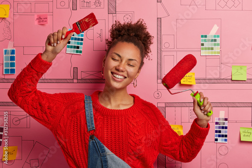 Satisfied relaxed ethnic woman raises both hands with painting tools, smiles broadly, wears red knitted sweater, does repairing inside of house, dances against room sketches, works on designing photo