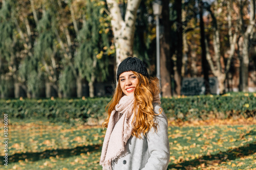 Blonde girl with dark blue cap, gray coat and pink scarf smiles as she walks through a park