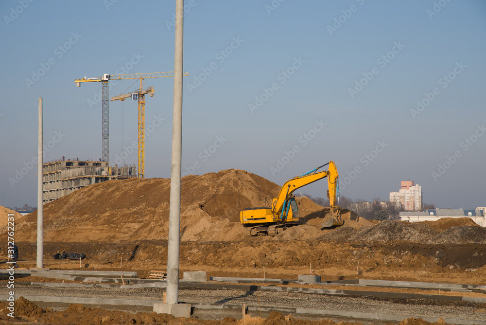 Excavator at a construction site on a background of a construction cranes and building. Backhoe dig the ground for the foundation, laying storm sewer pipes. Installation of water main systems.