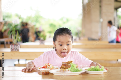 Asian child or kid girl wow and stunned surprise with enjoy eating grilled chicken and papaya salad for Thai food and appetizing on table with stick out tongue and funny face for lunch in restaurant
