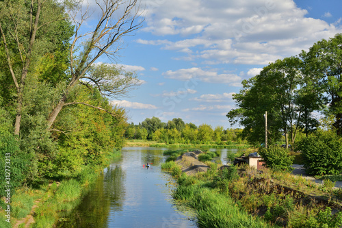 Ujście kanału wodnego do rzeki wśród drzew i chmur na niebie w letni dzień.