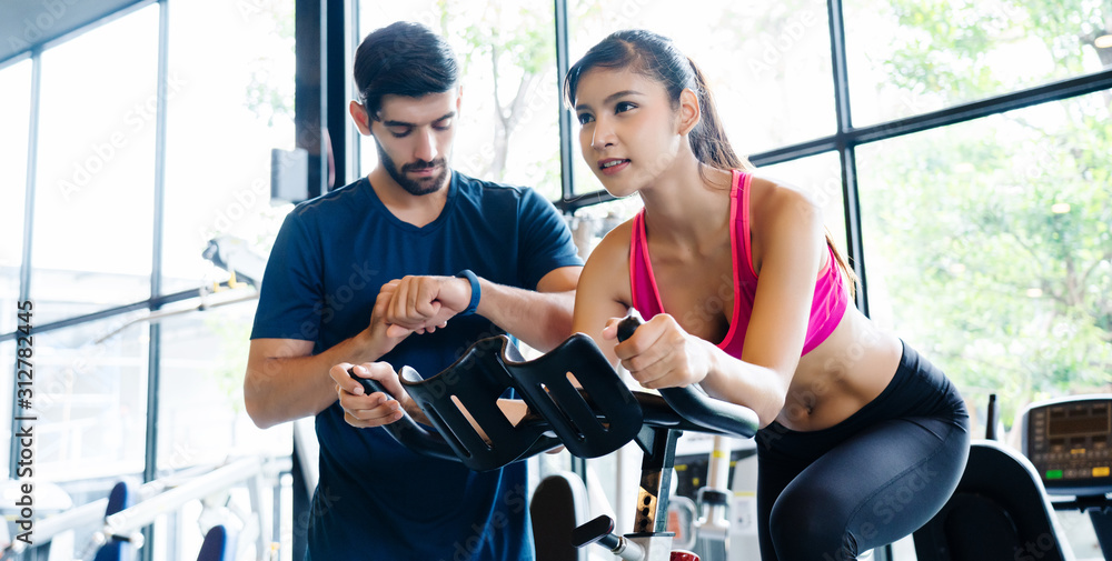 Fototapeta premium Asian Women wear sportswear, workout ride bicycles in gym, with the intention of health care. With a trainer to advise By cycling to make the body healthy With tight muscles And reduce the weight good