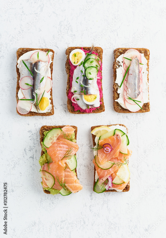Fototapeta Savory fish smorrebrod, set of five traditional Danish sandwiches. Black rye bread with anchovy, beetroot, radish, eggs, salmon, cream cheese, cucumber, avocado on grey white stone table, top view