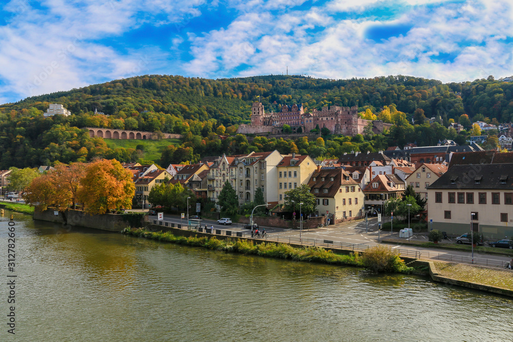 Lovely riverside view of the old town of Heidelberg, Germany and the famous Heidelberg Castle, a ruin and landmark on the northern part of the Königstuhl hillside on a nice autumn day with a blue sky.