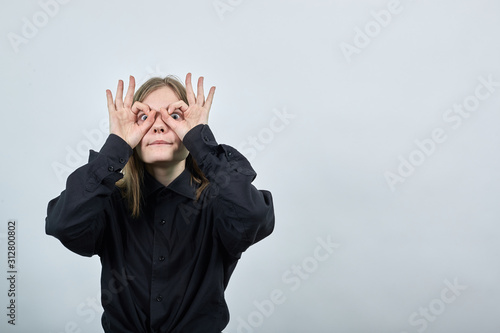 Crazy caucasian young woman in fashion black shirt isolated on gray background in studio doing okay gesture, glasses near eyes. People sincere emotions, lifestyle concept.