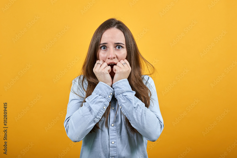 Scared attractive caucasian woman wearing fashion shirt isolated on orange background in studio biting nails, keeping fists near mouth. People emotions, lifestyle concept. Scared attractive caucasian woman wearing fashion shirt isolated on orange background in studio biting nails, keeping fists near mouth. People emotions, lifestyle concept.