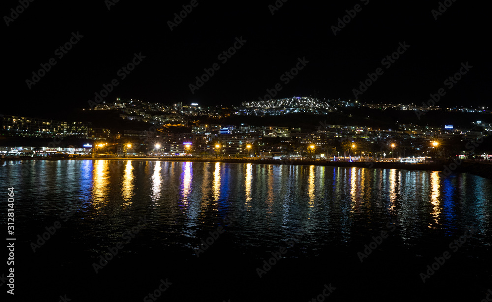 Night photography of the city with blue and yellow lights reflecting in the ocean. Puerto Rico, Gran Canaria, Spain.
