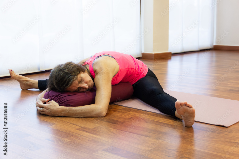 Middle aged woman in dragonfly pose lying forward on bolster. Young lady with open legs in straddle asana at bright studio. Yin yoga class concept Middle aged woman in dragonfly pose lying forward on bolster. Young lady with open legs in straddle asana at bright studio. Yin yoga class concept