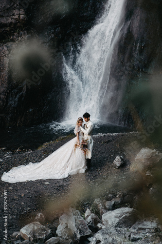 noise effect, selective focus: incredibly enamored brides hugging, kissing and posing for a photo on the incredible rocky mountains background with a big waterfall