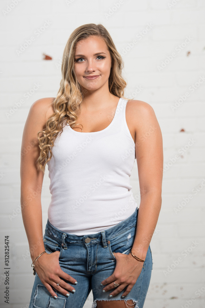 Sexy young blonde woman poses in studio wearing white tank top and ripped  blue jeans Stock Photo | Adobe Stock