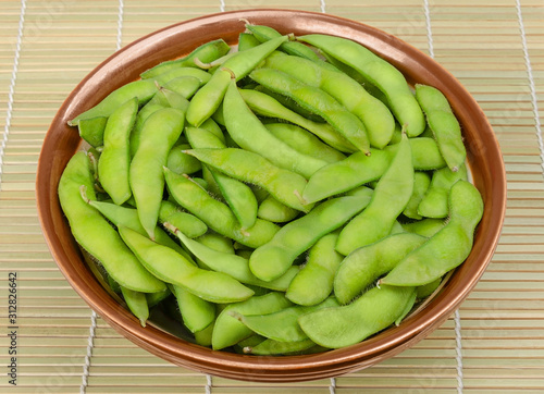 Boiled edamame in a brown bowl on a bamboo mat. Green soybeans, also maodou, cooked in saltwater. A side dish and tasty snack. Soy beans, Glycine max., a rich on protein legume. Closeup, food photo.