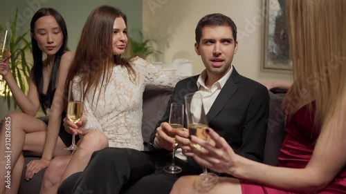 Handsome womanizer male sitting on sofa, talking and flirting with three beautiful young women over sparkling wine at party photo