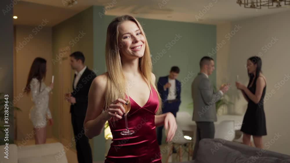 Attractive young woman in sexy red dress dancing with flute of champagne and looking at camera at house party, young people talking and relaxing in background Attractive young woman in sexy red dress dancing with flute of champagne and looking at camera at house party, young people talking and relaxing in background