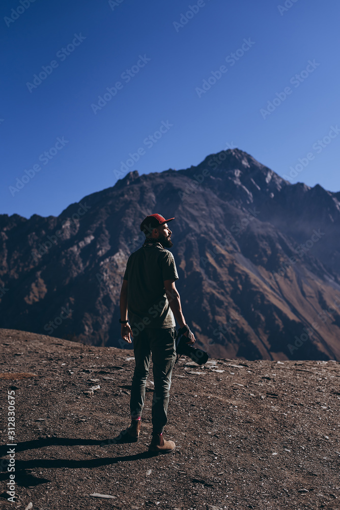 stylish bearded man with camera in hands on top of rocky Mountains: Outdoor photographer in caucasus mountains