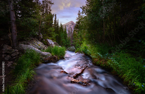 Long exposure of a river in the Rocky Mountains at sunset. There are pine trees lining the river and a mountain peak can be seen in the distance.