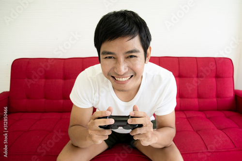 Asian young man sitting on a red sofa and concentrate on playing a video game. Man addicted playing a video game.