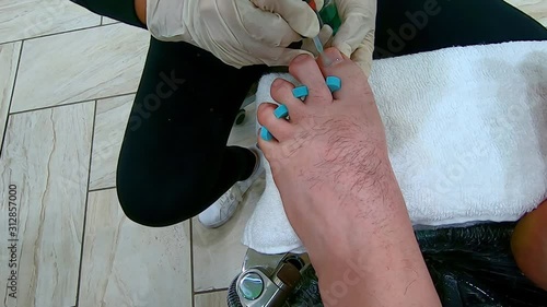 Professional Pedicurist wearing rubber gloves painting the left toe nails of a man's toes with blue rubber tool photo