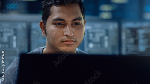 Portrait of a Smart and Handsome IT Specialist Working on a Personal Computer. In the Background Unfocused Personal Computers with Screens in Technologically Advanced Information and Data Center