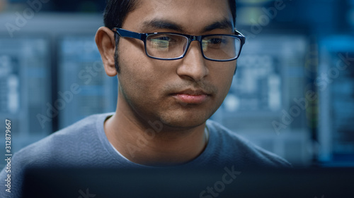 Portrait of a Smart and Handsome Software Developer Wearing Glasses Working on a Personal Computer, Smiles at the Camera. In the Background Unfocused Personal Computers with Screens in Technical Room © Gorodenkoff