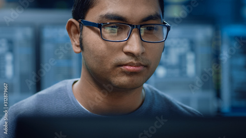 Portrait of a Smart and Handsome Software Developer Wearing Glasses Working on a Personal Computer, Smiles at the Camera. In the Background Unfocused Personal Computers with Screens in Technical Room