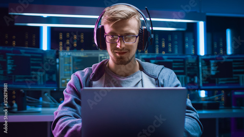 Portrait of Software Developer / Hacker / Gamer Wearing Glasses and Headset Sitting at His Desk and Working / Playing on Laptop. In the Background Dark High Tech Environment with Multiple Displays. photo