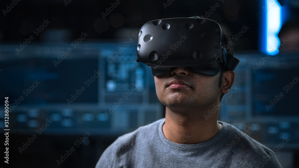 Software Delevoper Wearing Virtual Reality Headset on His Head, Developing and Programming VR Game or Application. In the Background Technology Developing Studio with Computers and Monitors Software Delevoper Wearing Virtual Reality Headset on His Head, Developing and Programming VR Game or Application. In the Background Technology Developing Studio with Computers and Monitors