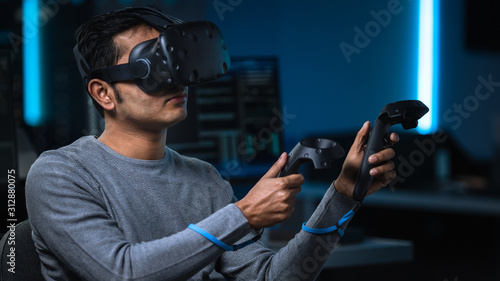 Portrait of Software Delevoper Wearing Virtual Reality Headset Using Controllers to Develop and Program VR Gaming and Applications. In Background Technology Developing Studio with Computer Monitors