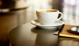 Close-up white cup of coffee with mobile phone and notebook on desk.  Background vintage light