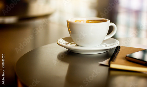 Close-up white cup of coffee with mobile phone and notebook on desk. Background vintage light
