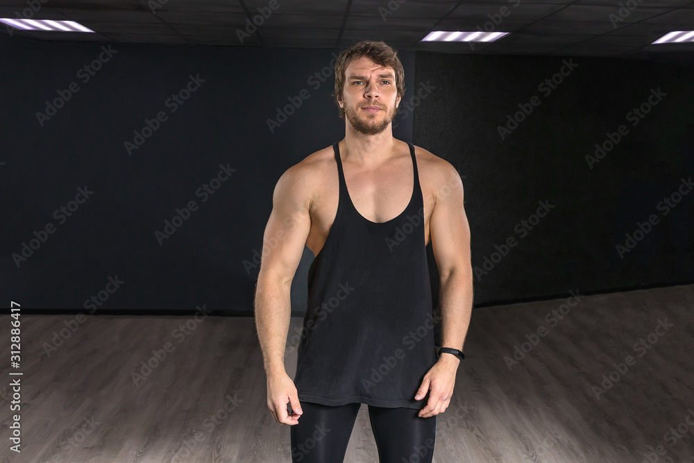 Obraz premium Muscular strong athletic guy in a black tank top standing posing near dark wall. Concept sport photo with copy space